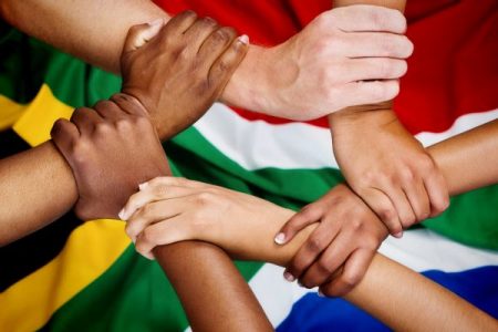 South African Flag in background with arms and hands of people with different skin colours all making a unified sign with hands grasping writs of each other